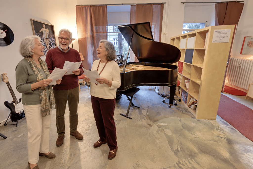 KI-generiertes Symbolbild: Drei Seniorinnen und Senioren singen gemeinsam vor dem Flügel an seinem ursprünglichen Platz im Musikschulraum.