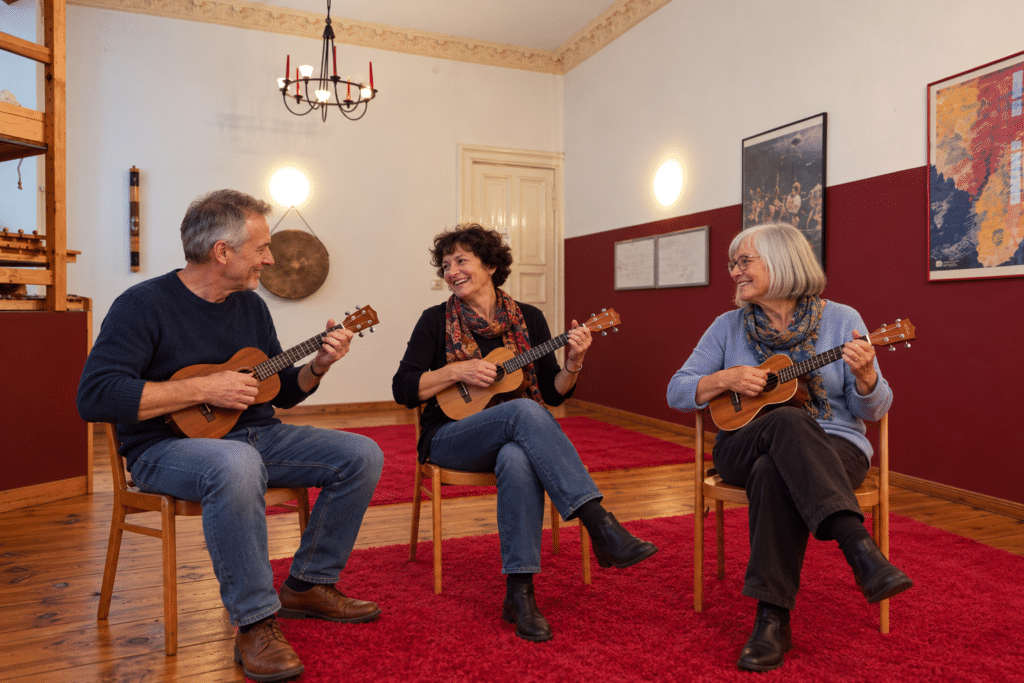 KI-generiertes Symbolbild: Drei Seniorinnen und Senioren spielen sitzend gemeinsam Ukulele im nachgebildeten weinroten Raum der moamusikschule.